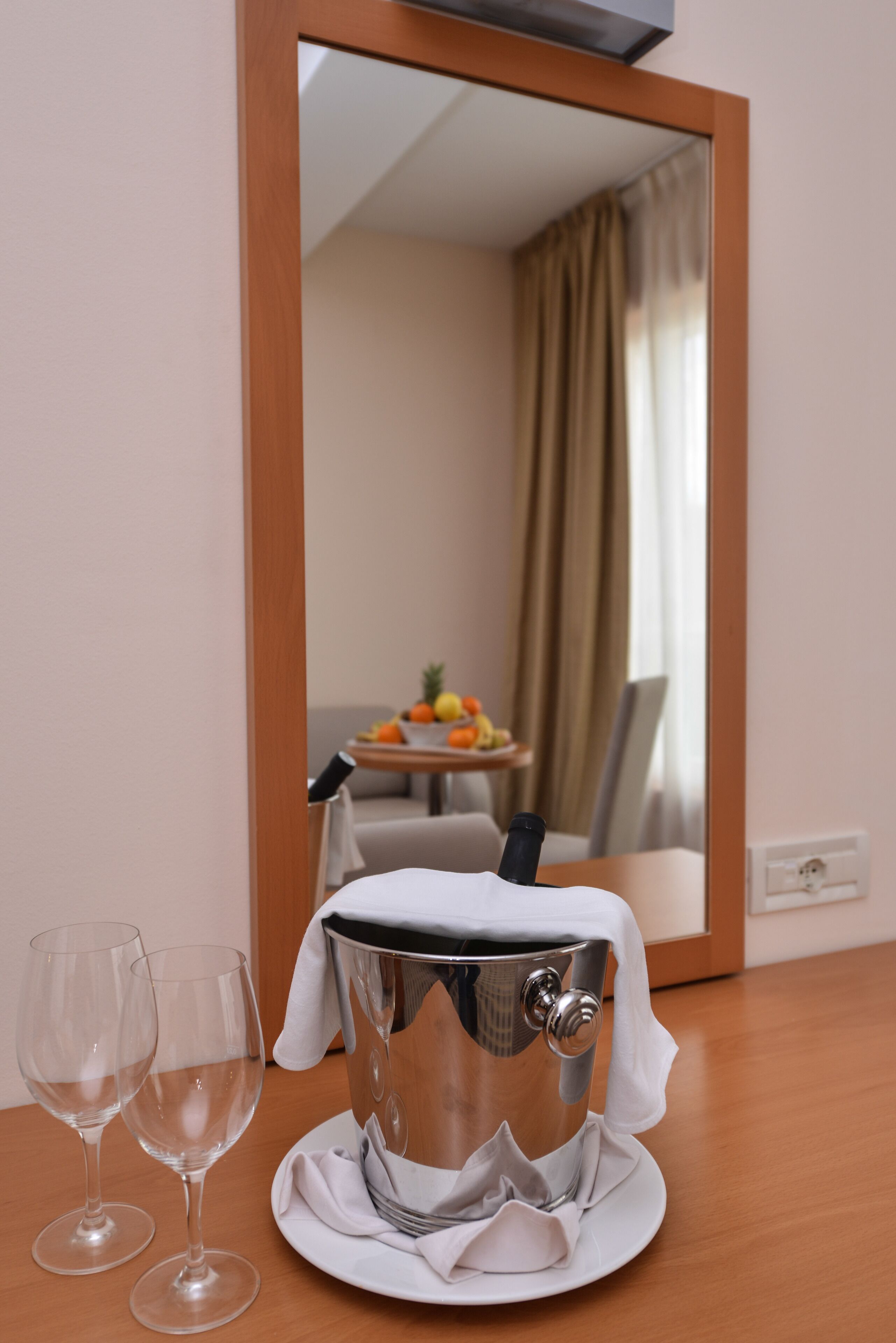Ein Hotelzimmer mit einem Holztisch, zwei leeren Weingläsern, einem silbernen Eimer mit Deckel und einem Spiegel, der einen Essbereich mit Obstschale und Vorhängen zeigt.