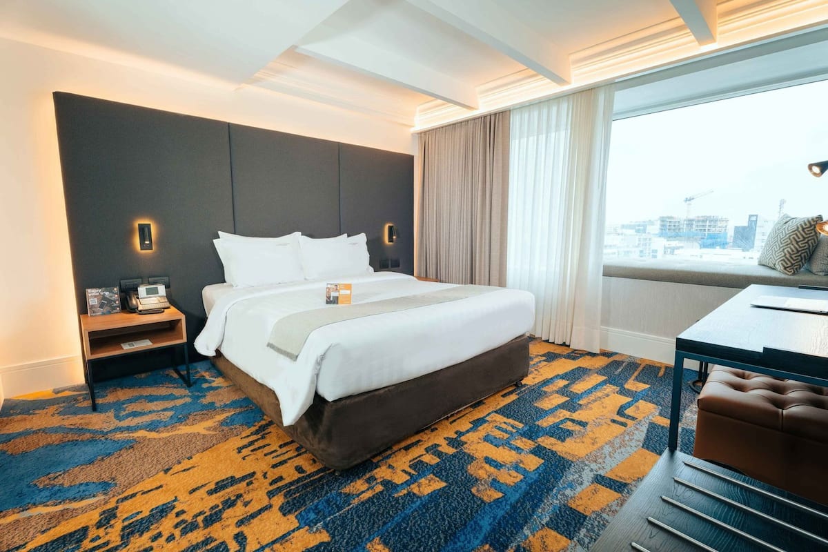 Une chambre d’hôtel avec un grand lit, un bureau avec une chaise, une table de chevet et une vue sur la ville par la fenêtre.