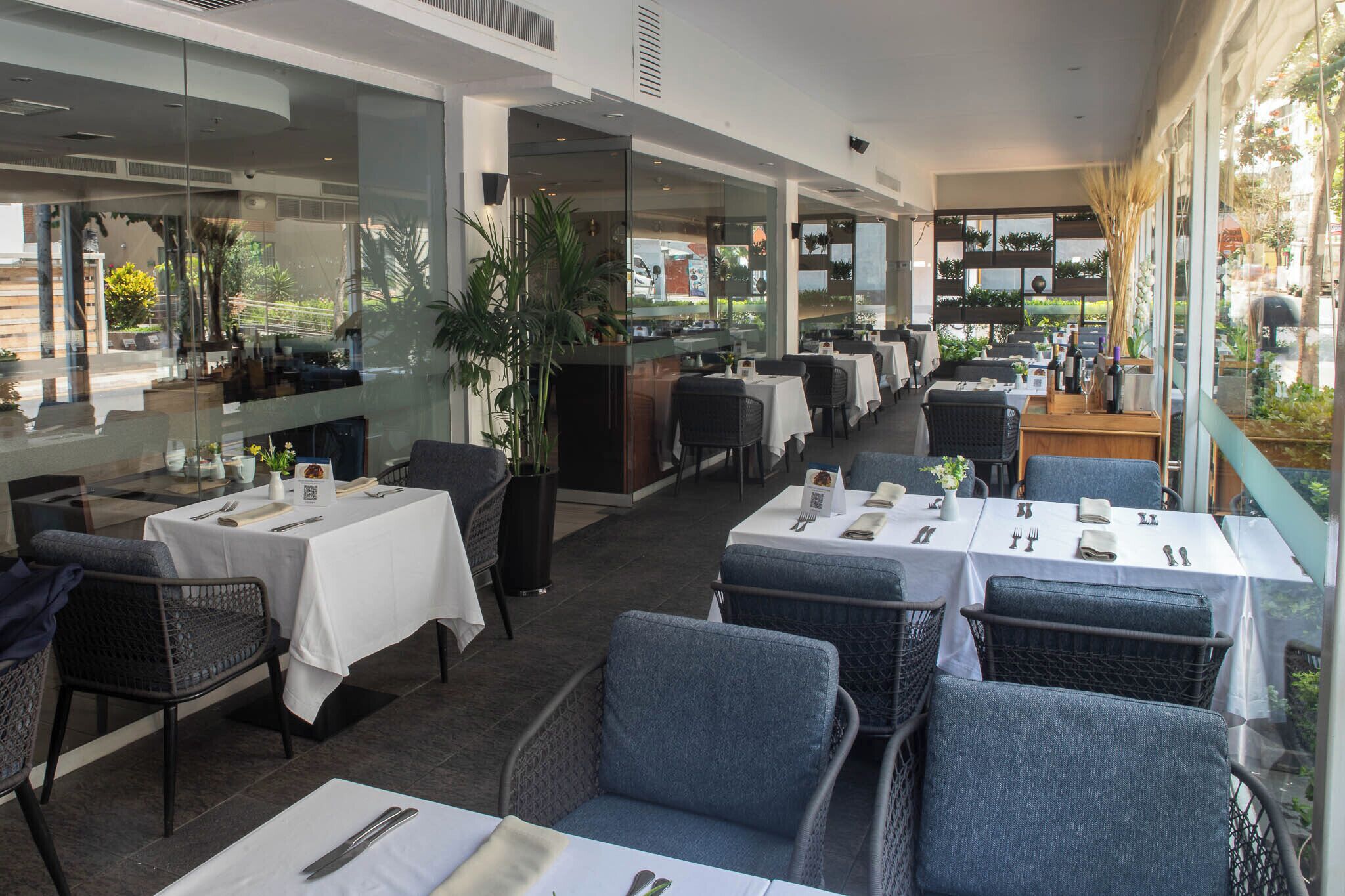 An outdoor dining area with tables and chairs, large windows, and potted plants.