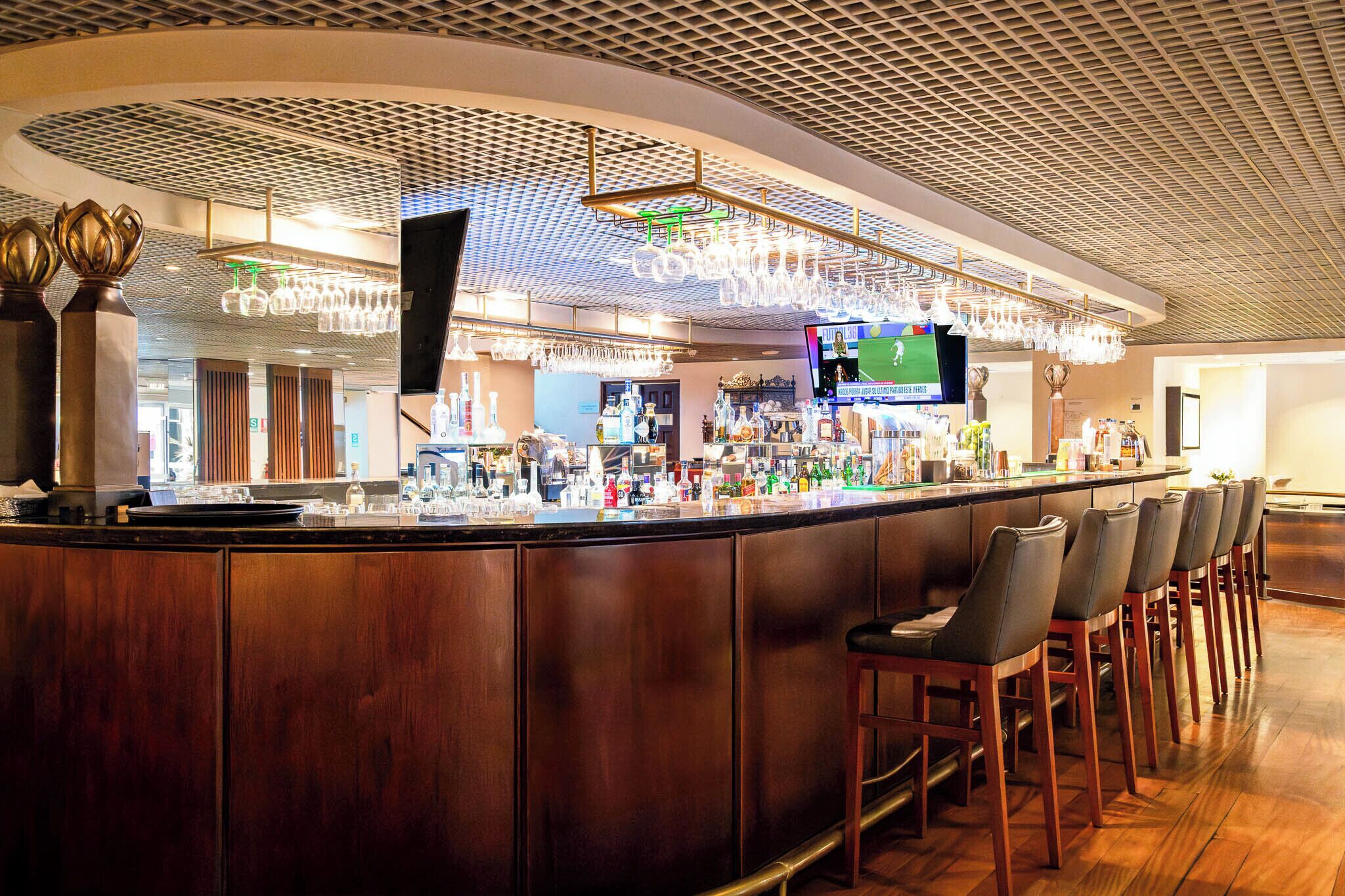 A bar area with a wooden counter, bar stools, and a television screen displaying a sports game.
