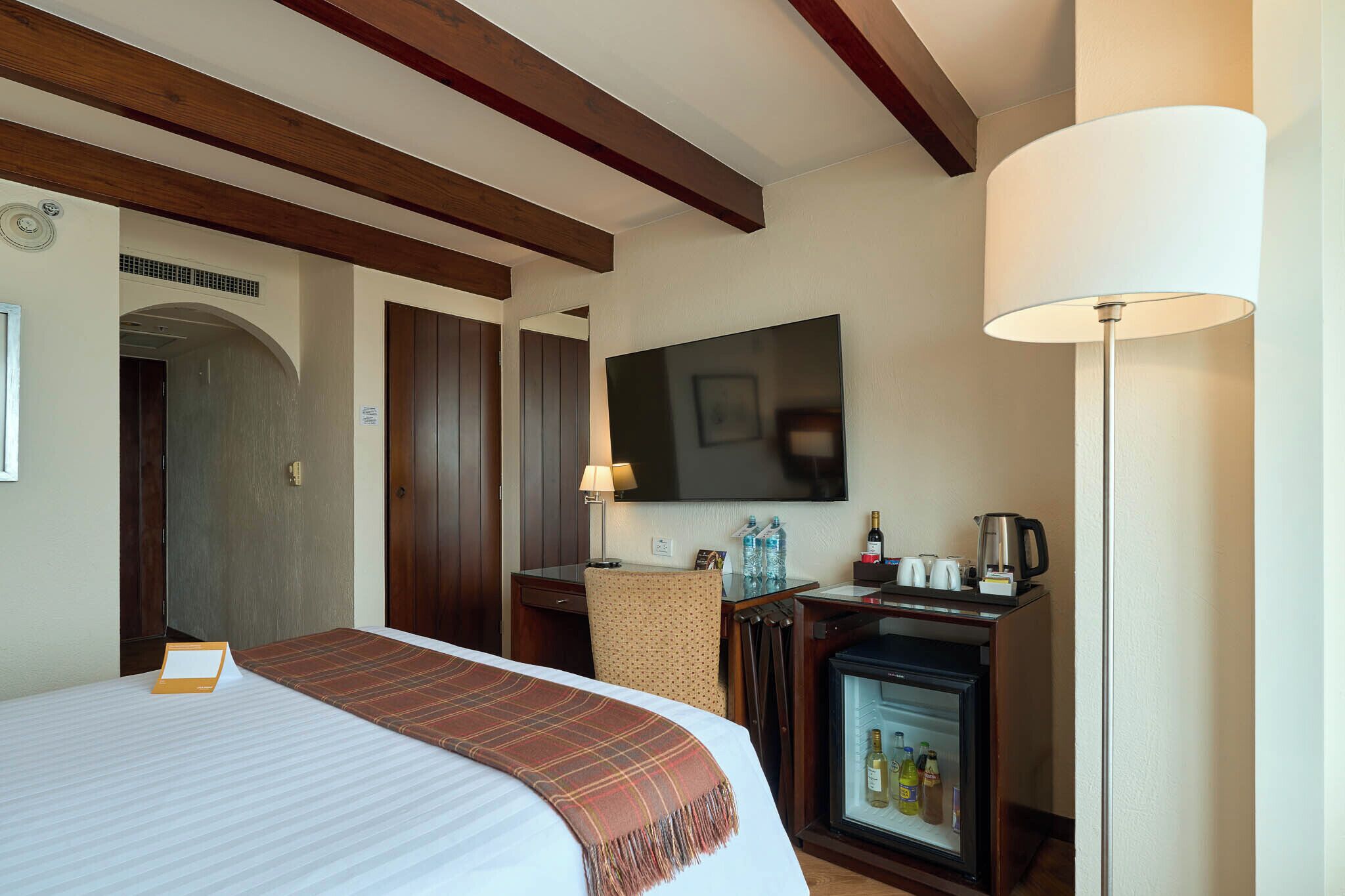 A hotel room with a bed, a desk with a lamp, a television, a refrigerator, and a door leading to another room.