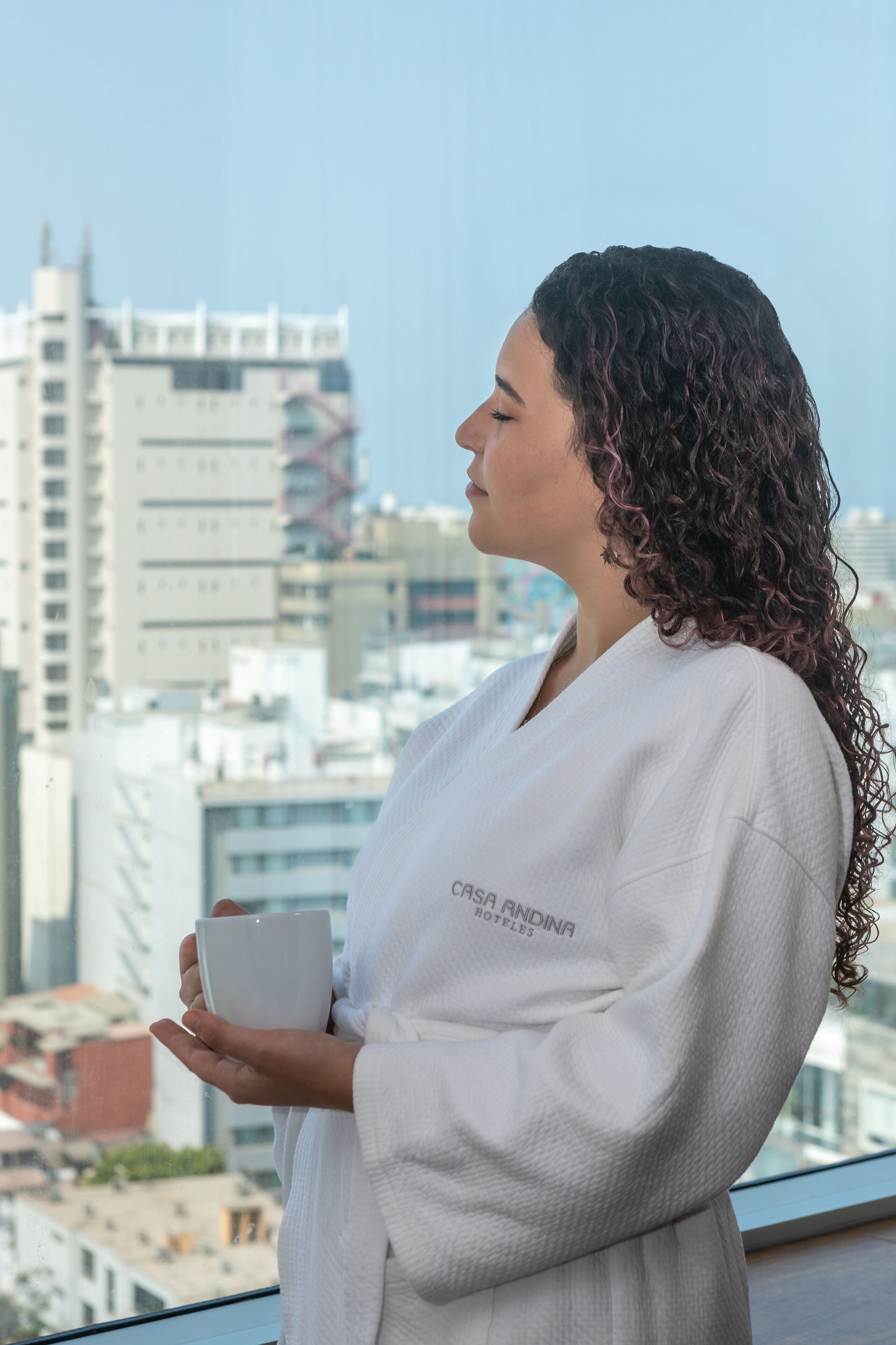 A person in a white robe holding a white mug, standing by a window with a cityscape view.