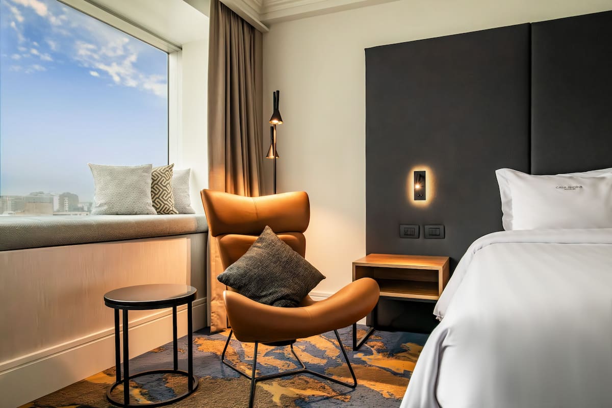 Une chambre d’hôtel avec un grand lit, un coin salon comprenant un fauteuil et une petite table, une table de chevet et une vue sur la ville par la fenêtre.