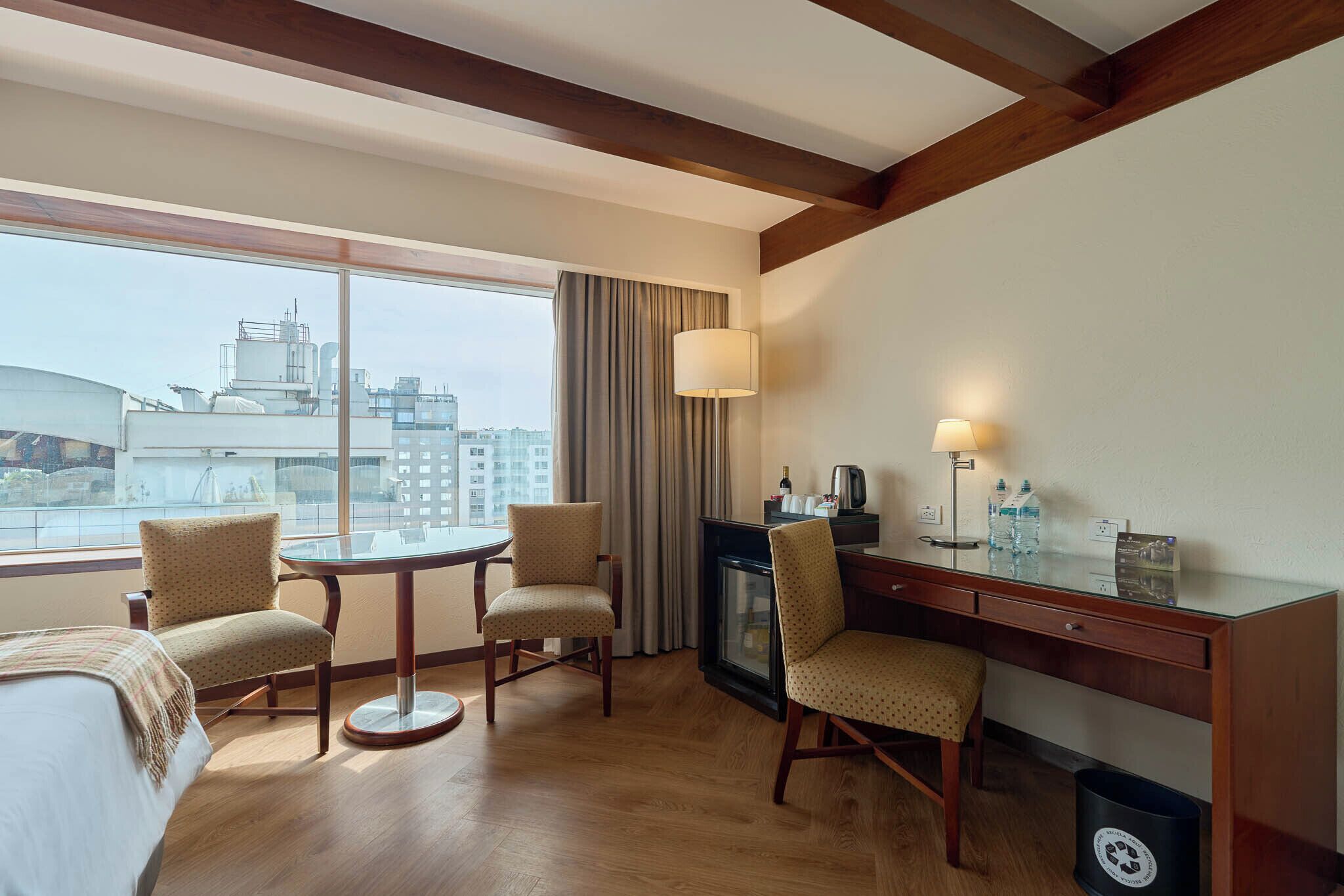 Habitación de hotel con un ventanal grande, una mesa redonda, dos sillas, un escritorio con lámpara y un cesto de basura.