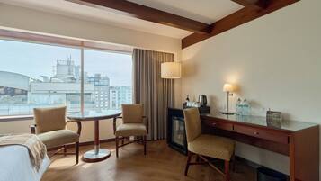 Habitación de hotel con un ventanal grande, una mesa redonda, dos sillas, un escritorio con lámpara y un cesto de basura.