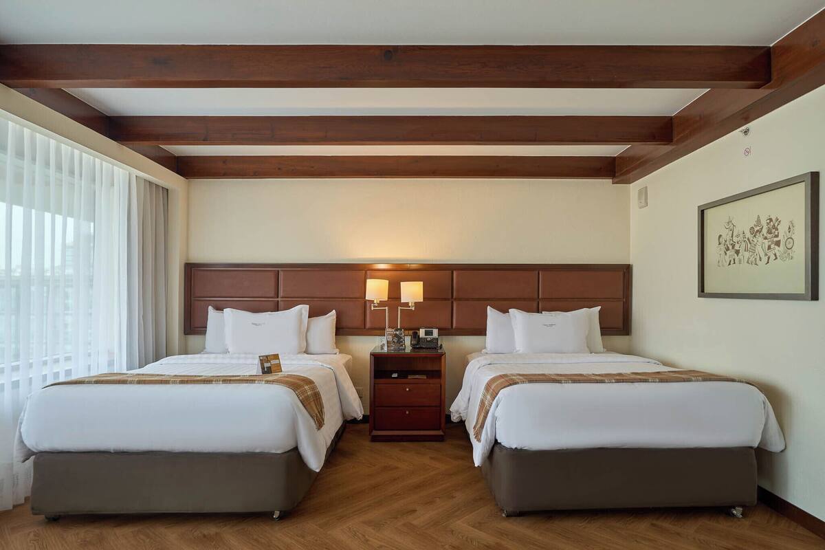 Une chambre d’hôtel avec deux lits, des poutres en bois au plafond, une table de chevet avec des lampes et un tableau encadré au mur.