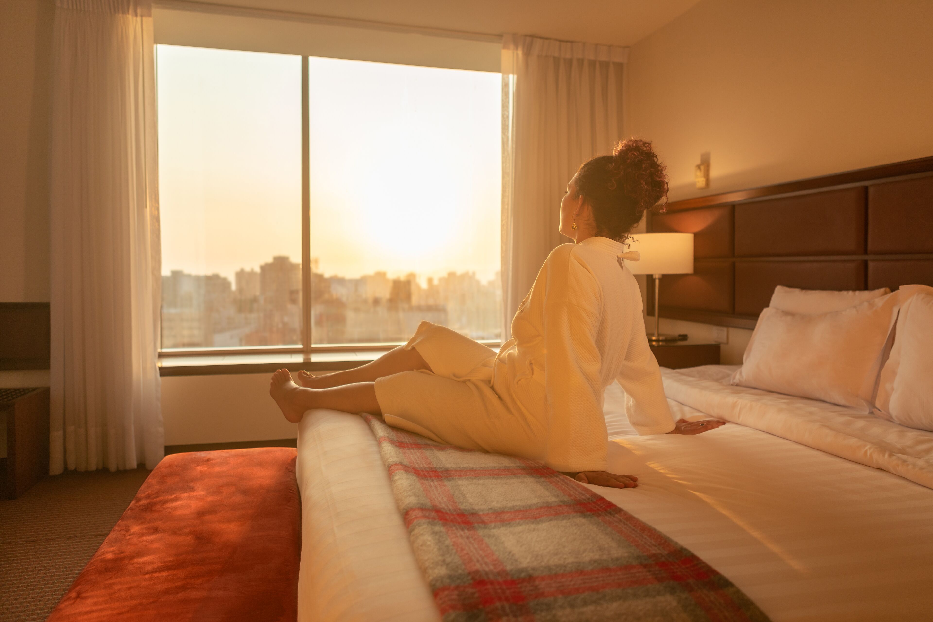 Una habitación de hotel con un ventanal grande, una cama con manta escocesa y una mesita de noche con lámpara.