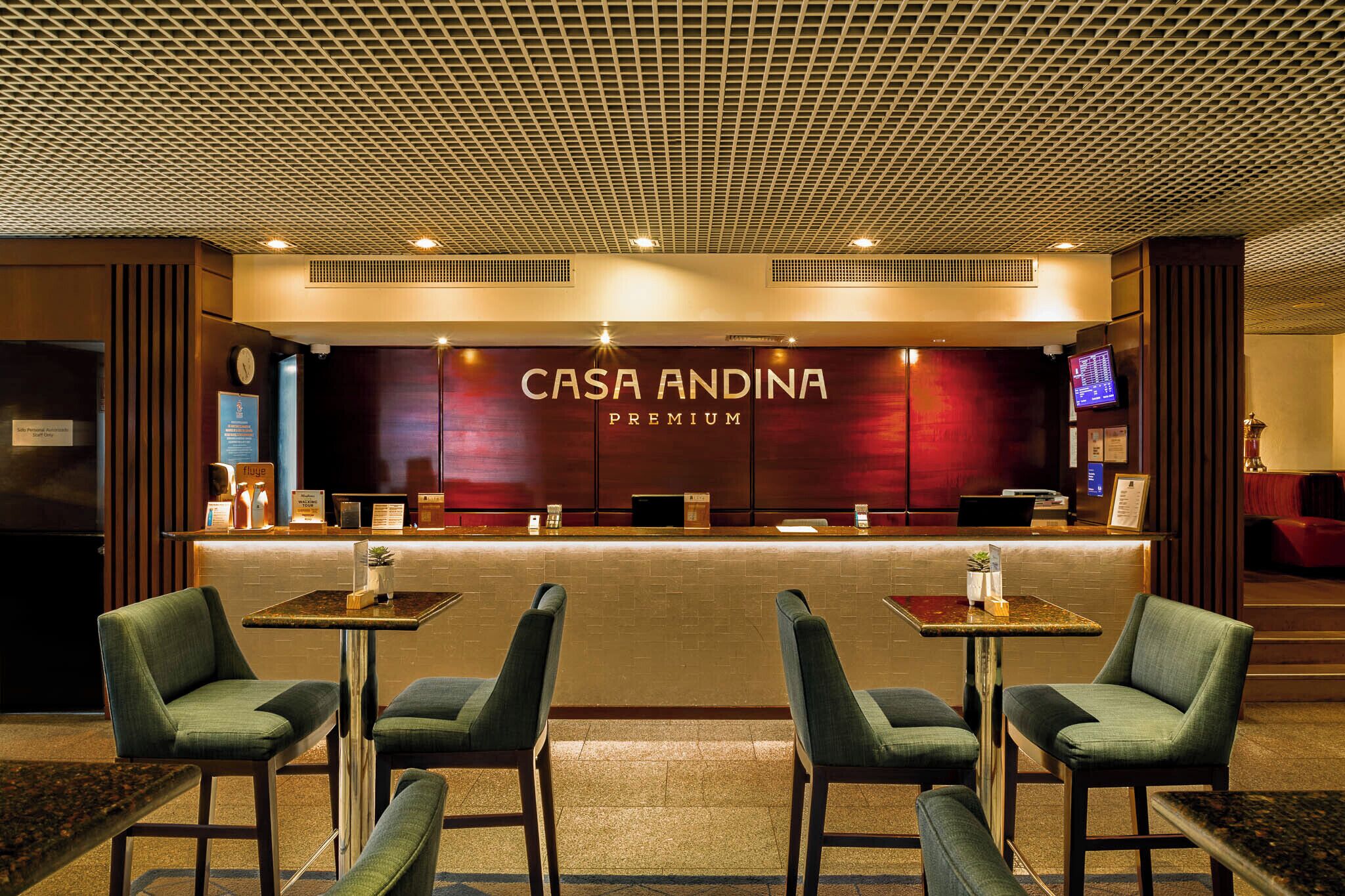 A hotel reception area with a 'CASA ANDINA PREMIUM' sign, a reception desk, seating area with tables and chairs, and a digital screen displaying information.