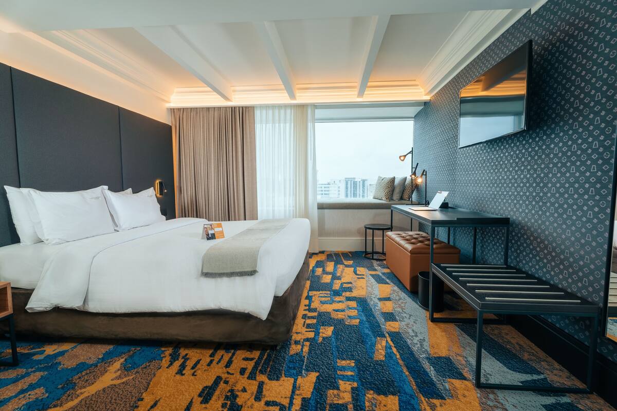 Une chambre d’hôtel avec un grand lit, un bureau, une chaise, un téléviseur fixé au mur et une vue sur la ville par la fenêtre.