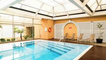 Una piscina interior con paredes de vidrio transparente, suelo de madera y techo con tragaluces.