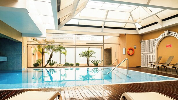 Una piscina interior con paredes de vidrio transparente, plataforma de madera y sillas de descanso.