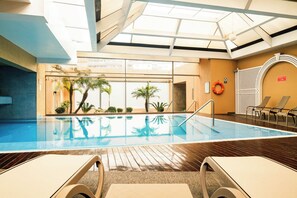Una piscina interior con paredes de vidrio transparente, plataforma de madera y sillas de descanso.