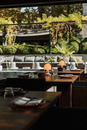 An outdoor dining area with tables set for two, featuring candlelit lamps and a view of a lush garden. - Roomers Frankfurt Central, Autograph Collection (Frankfurt)