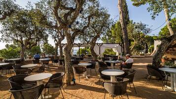 Zona d'esmorzar a l'aire lliure amb taules i cadires rodones, envoltada d'arbres i vistes al mar.