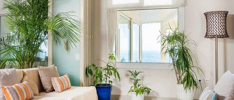 A living room with a sofa, two potted plants, a lamp, and a view of the sea through a window.