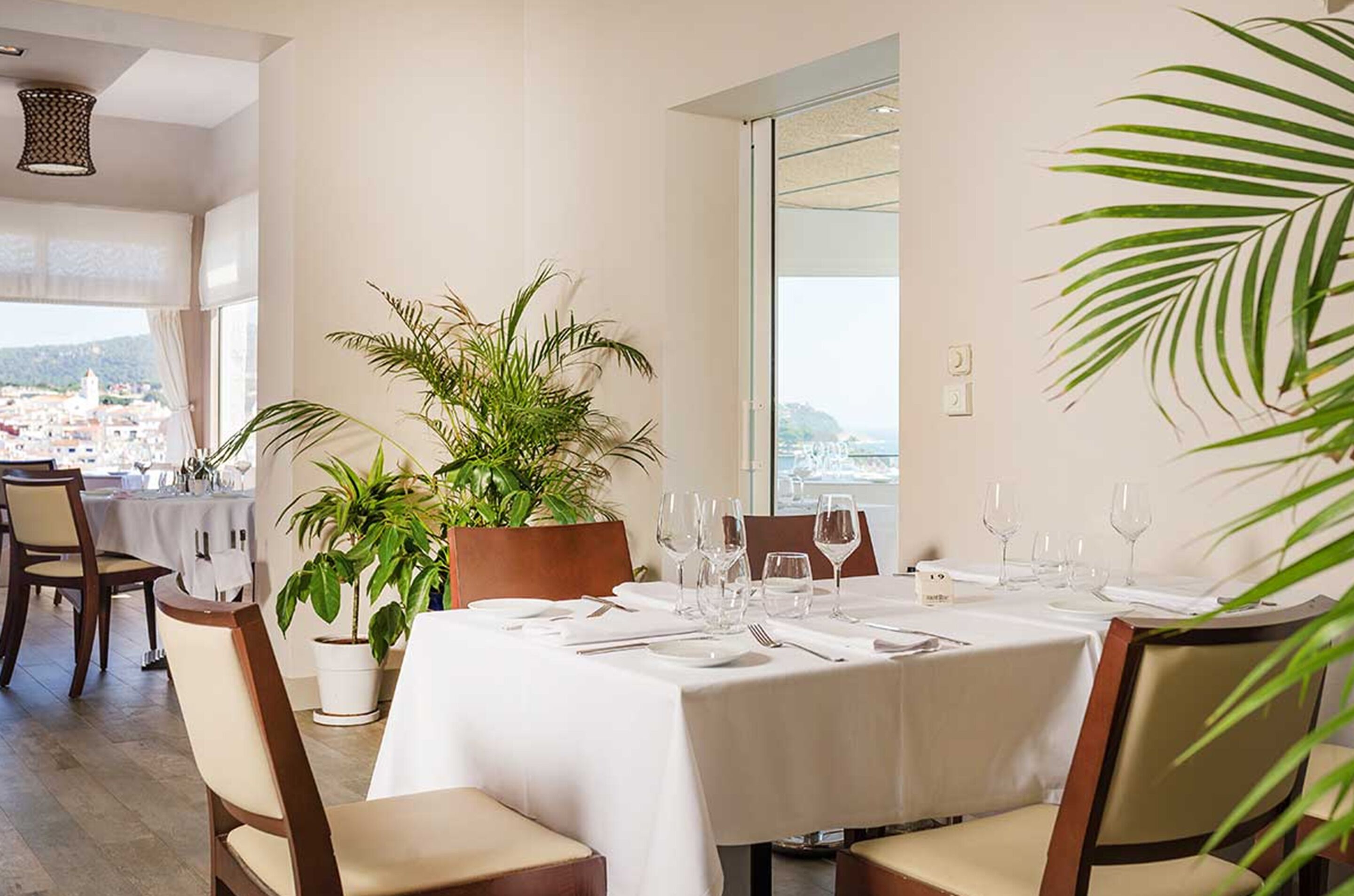 Un coin repas avec une table dressée pour quatre personnes, des plantes en pot et une vue sur le paysage urbain à travers de grandes fenêtres.