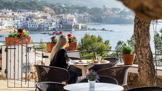 A terrace with wicker chairs and a table, overlooking a coastal town with buildings and the sea. - Hotel Sant Roc (Palafrugell)