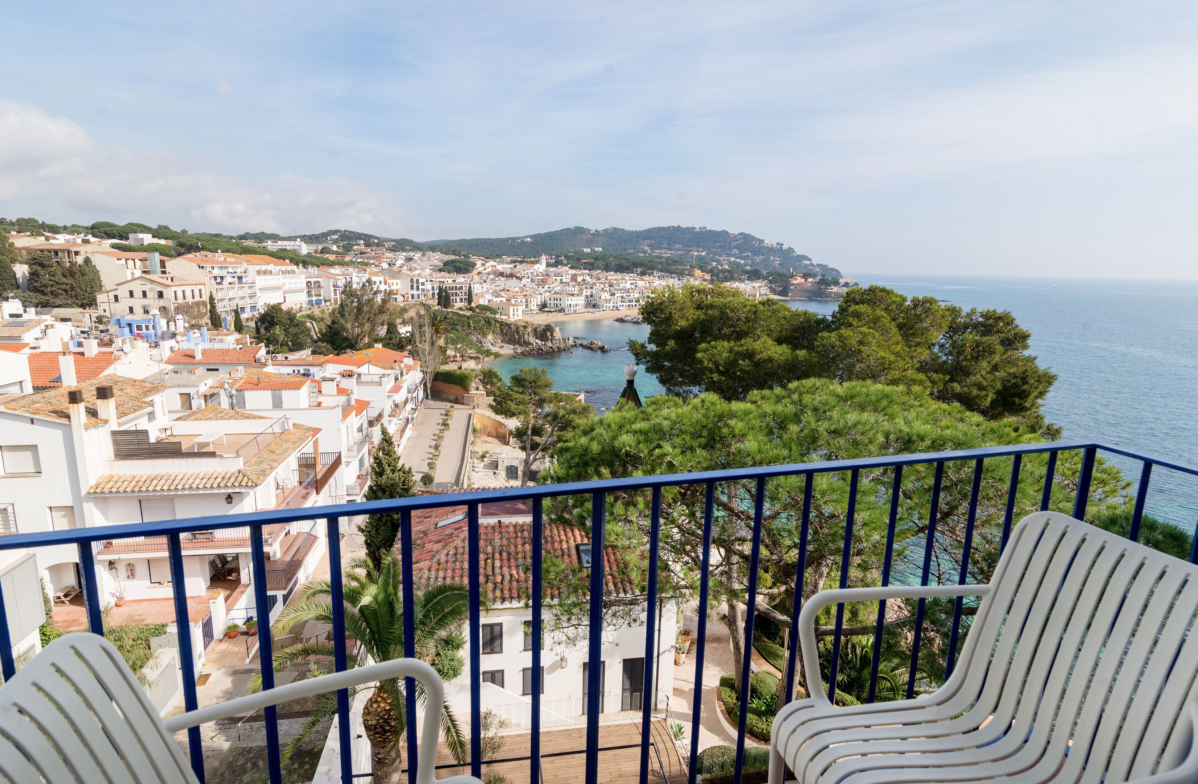 A balcony with two white chairs, overlooking a coastal town with buildings, trees, and a view of the sea.