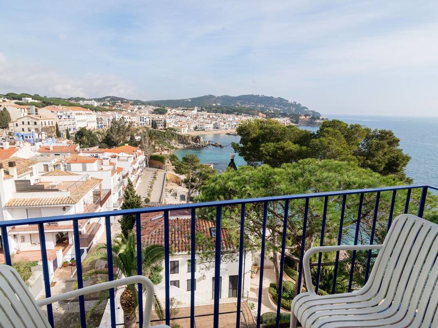 A balcony with two white chairs, overlooking a coastal town with buildings, trees, and a view of the sea.