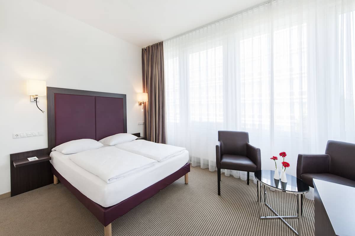 Et hotelværelse med seng, to stole, et lille bord og et vindue med gardiner.