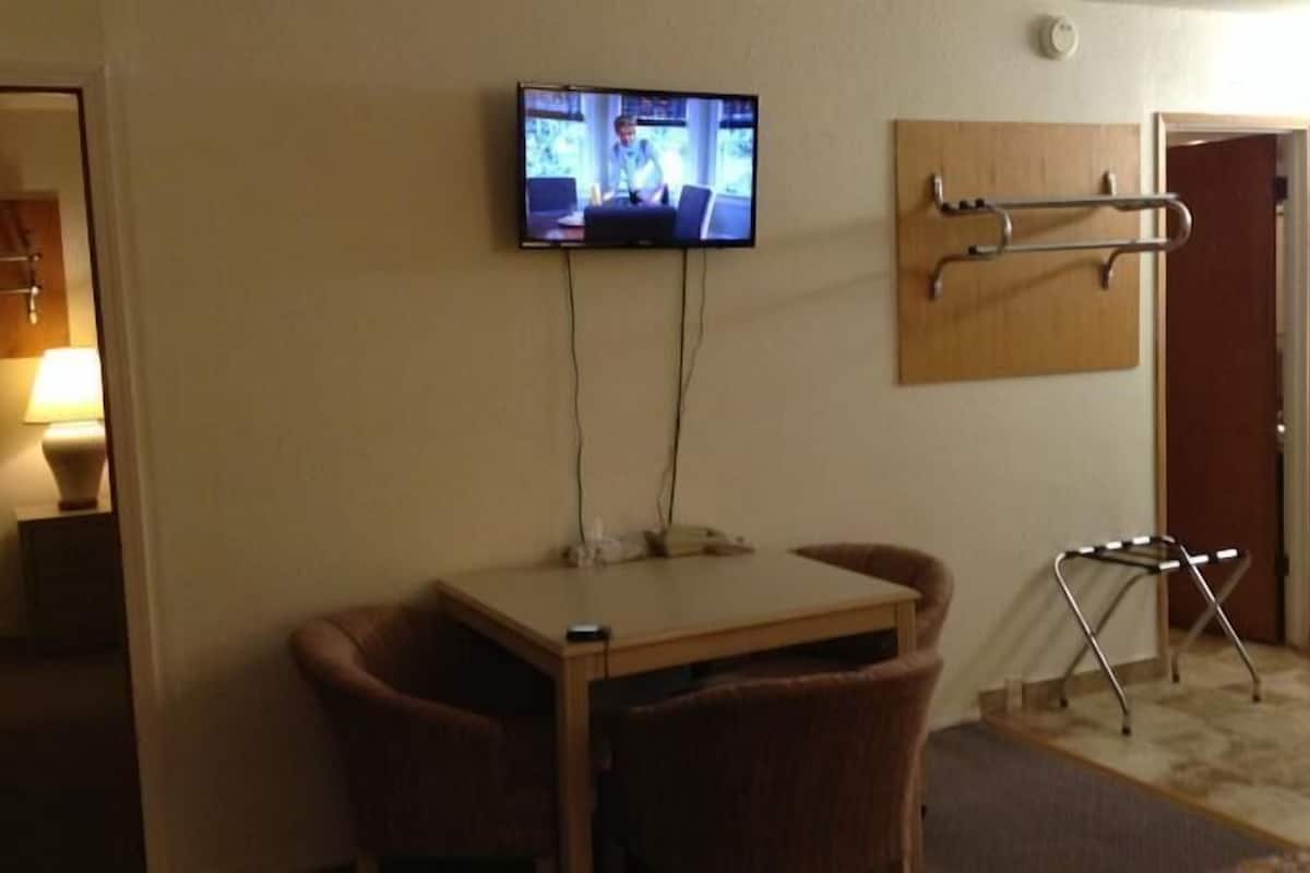 A small room with a television mounted on the wall, a table with two chairs, a small stool, and a door leading to a kitchenette.