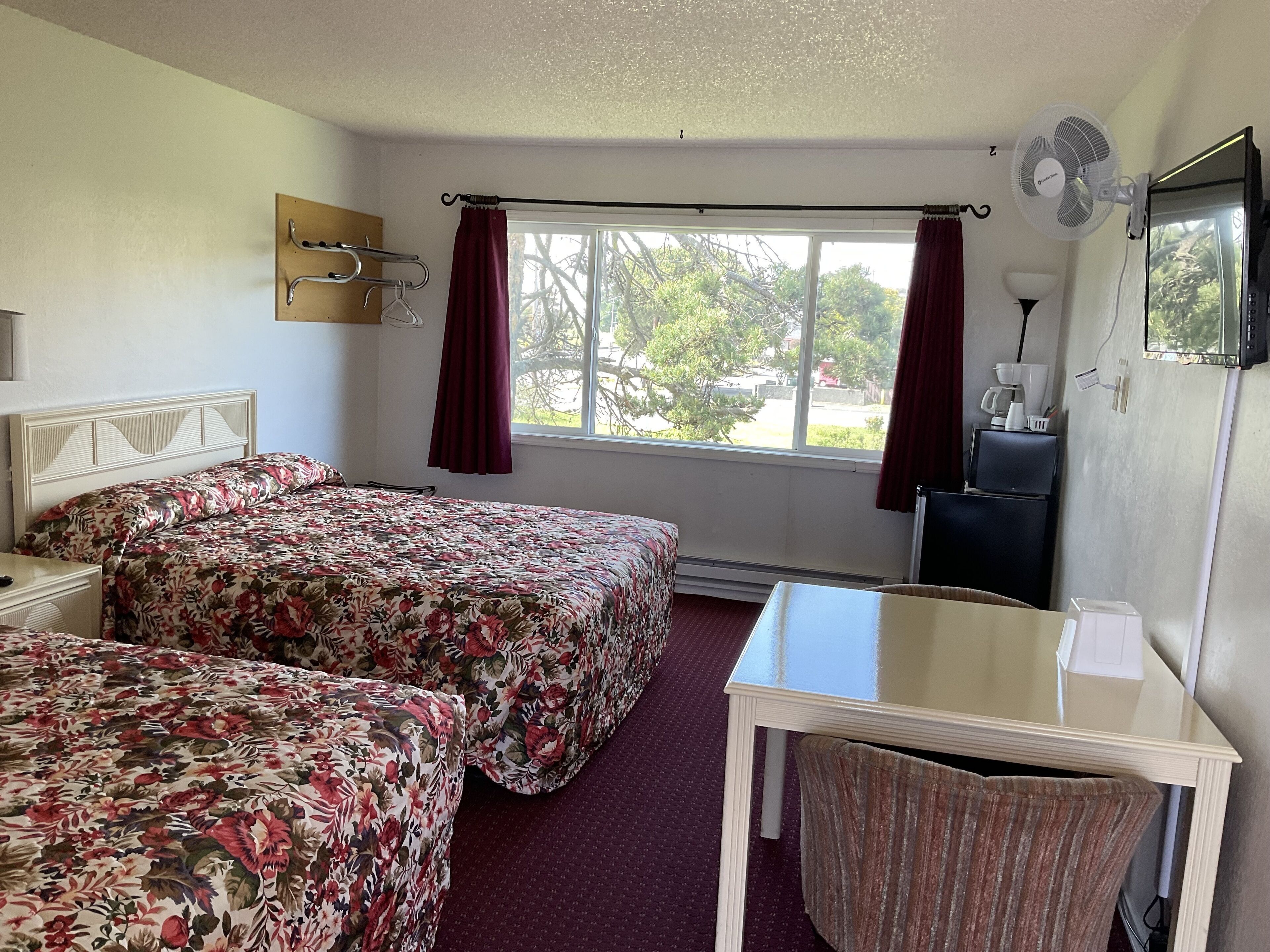 A hotel room with two beds, a small table, a chair, a desk, a mirror, a fan, and a window with curtains.