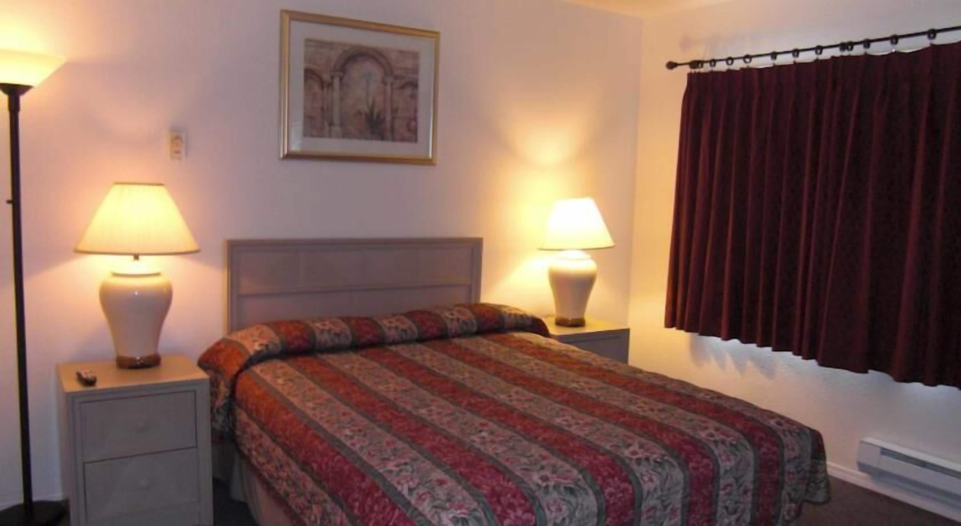 A neatly made bed with a patterned bedspread, two bedside lamps, a framed picture on the wall, and a window with a curtain.