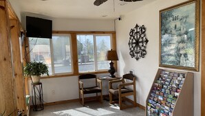 A room with a ceiling fan, a TV mounted on the wall, a table with chairs, a lamp, a window with a view of the street, and a display of magazines. - Front Street Inn (Crescent City)