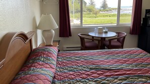 A hotel room with a bed, a table with chairs, a lamp, and a view of the beach through the window. - Front Street Inn (Crescent City)