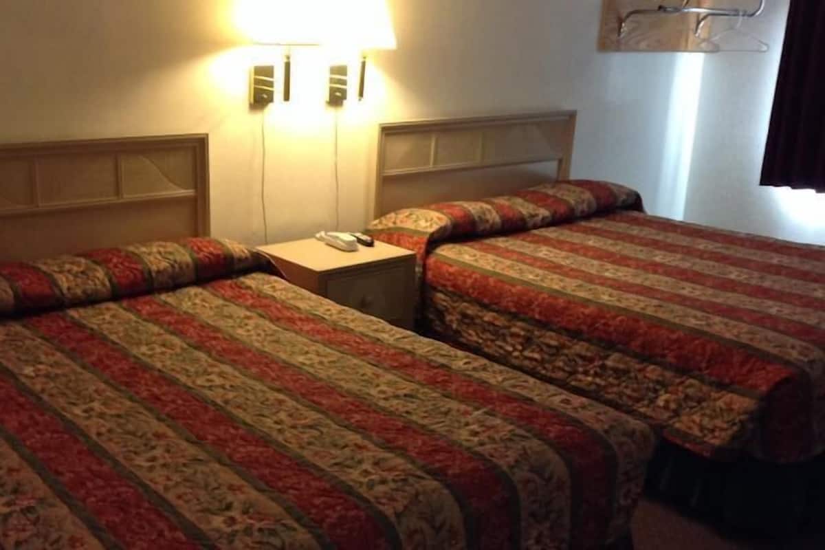 A hotel room with two beds, each with a patterned bedspread, a bedside table with a lamp, and a wall-mounted light fixture.