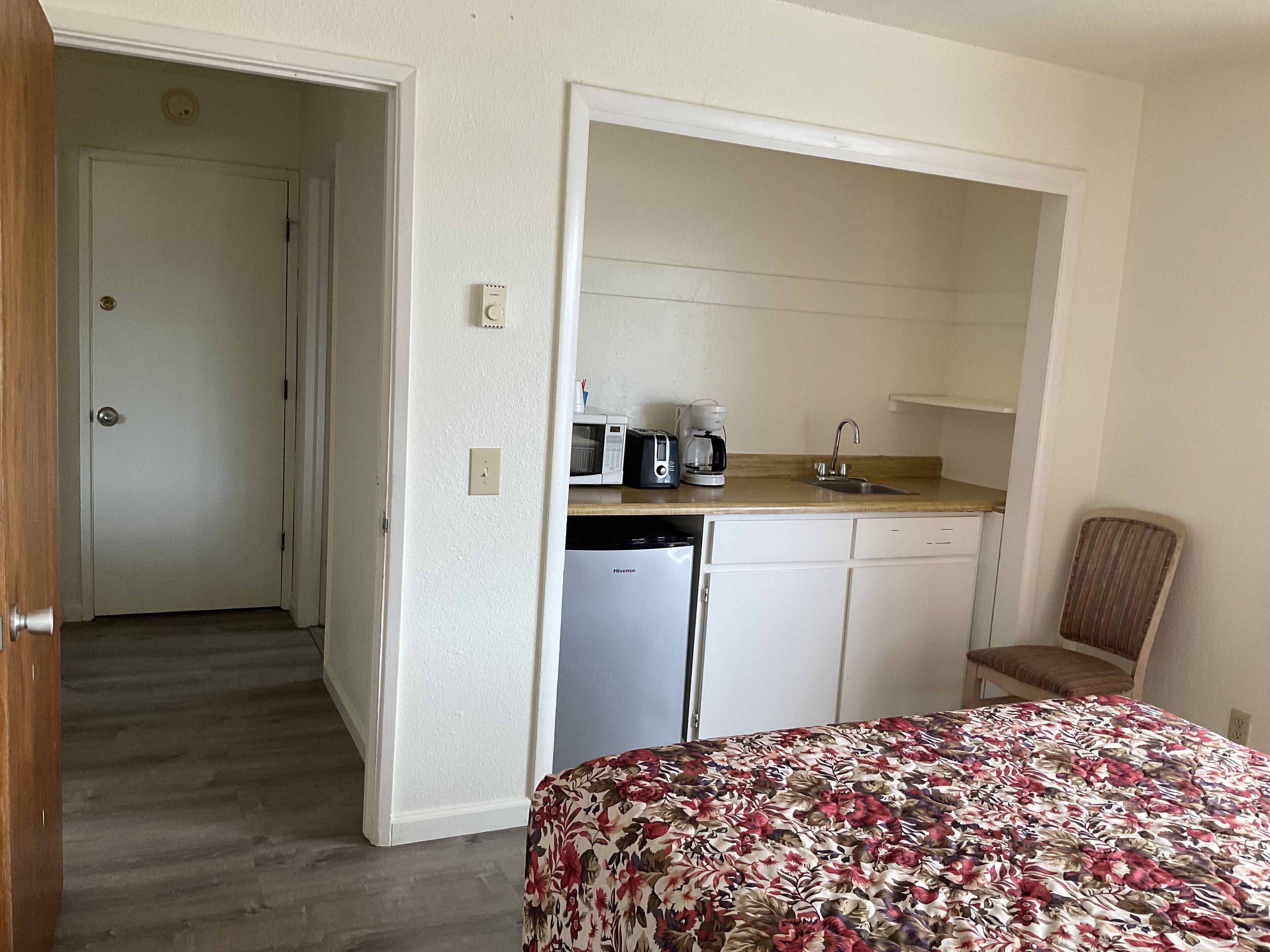 A compact hotel room with a bed, a kitchenette, a small sink, and a chair.