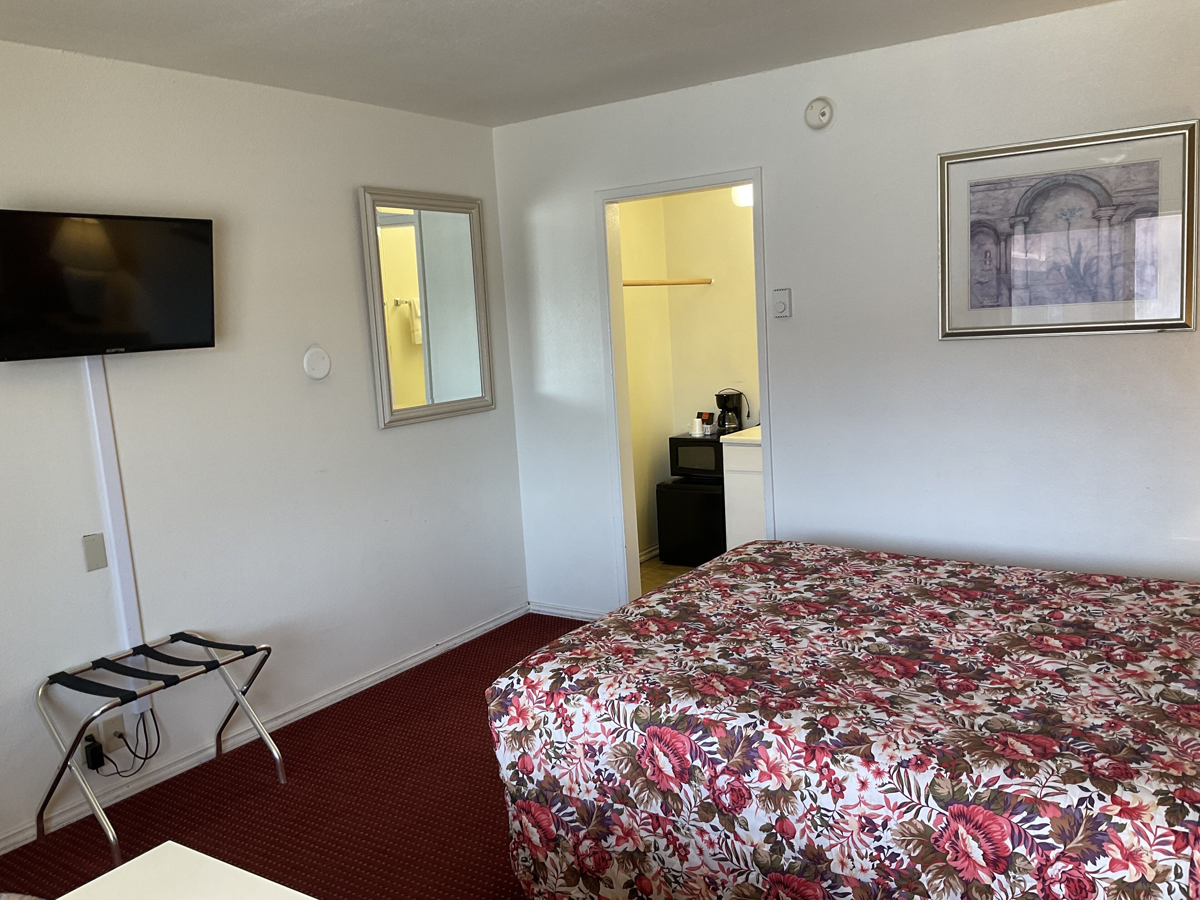 Habitación de hotel con cama, televisión, espejo, cuadro enmarco y pequeña mesa de trabajo con cafetera.