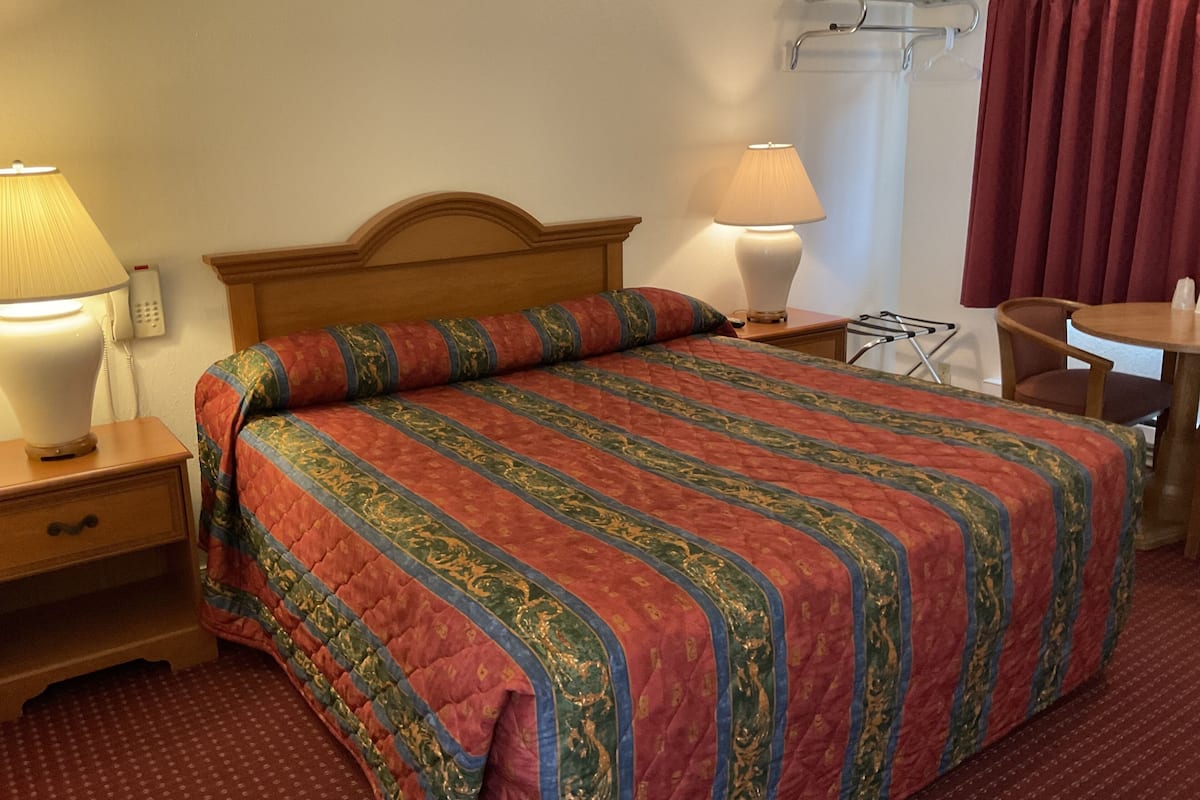 A hotel room with a wooden headboard bed, two bedside lamps, a small table, and a chair.