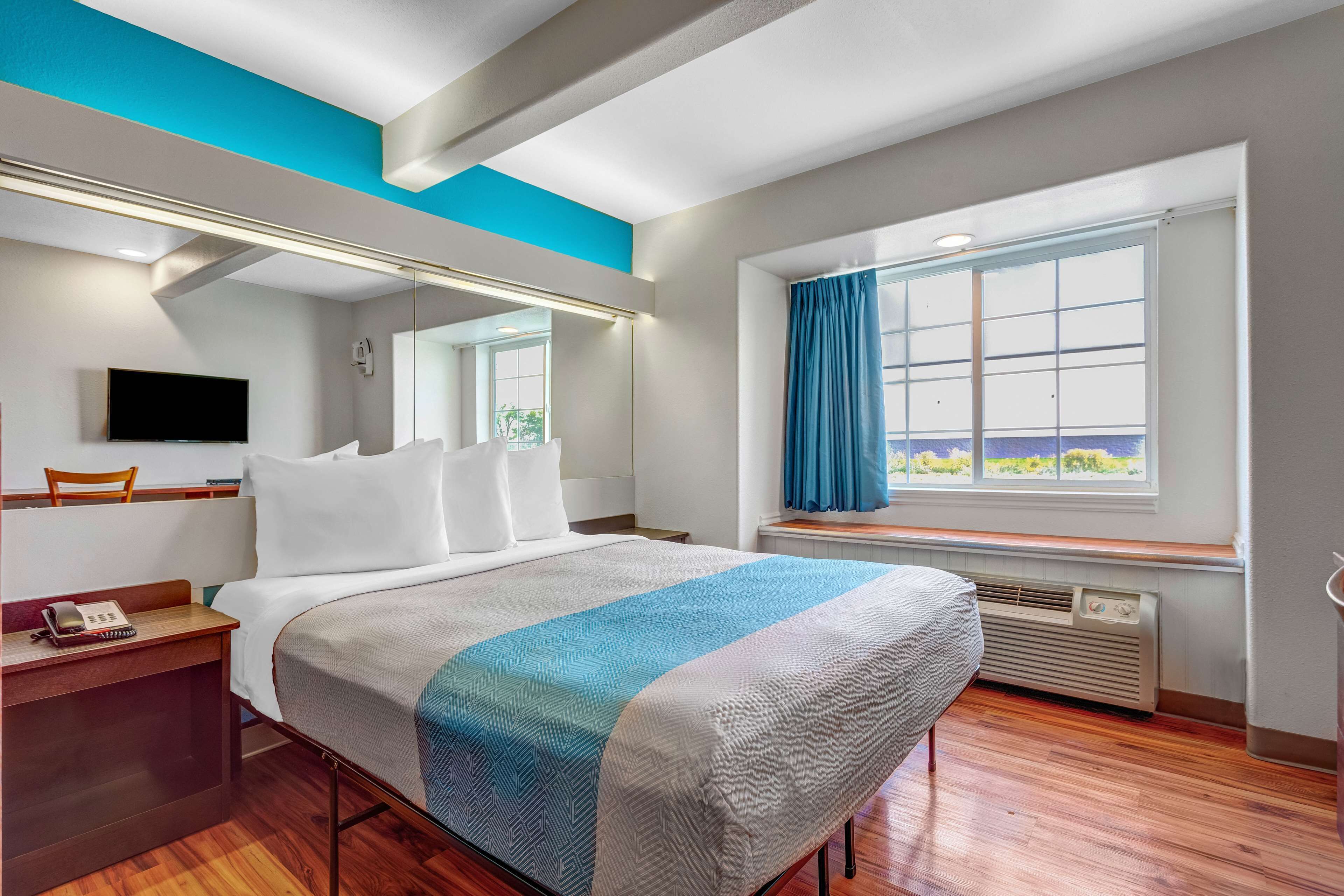 A hotel room with a bed, a desk, a chair, a TV, a window with curtains, and a view of the outdoors.