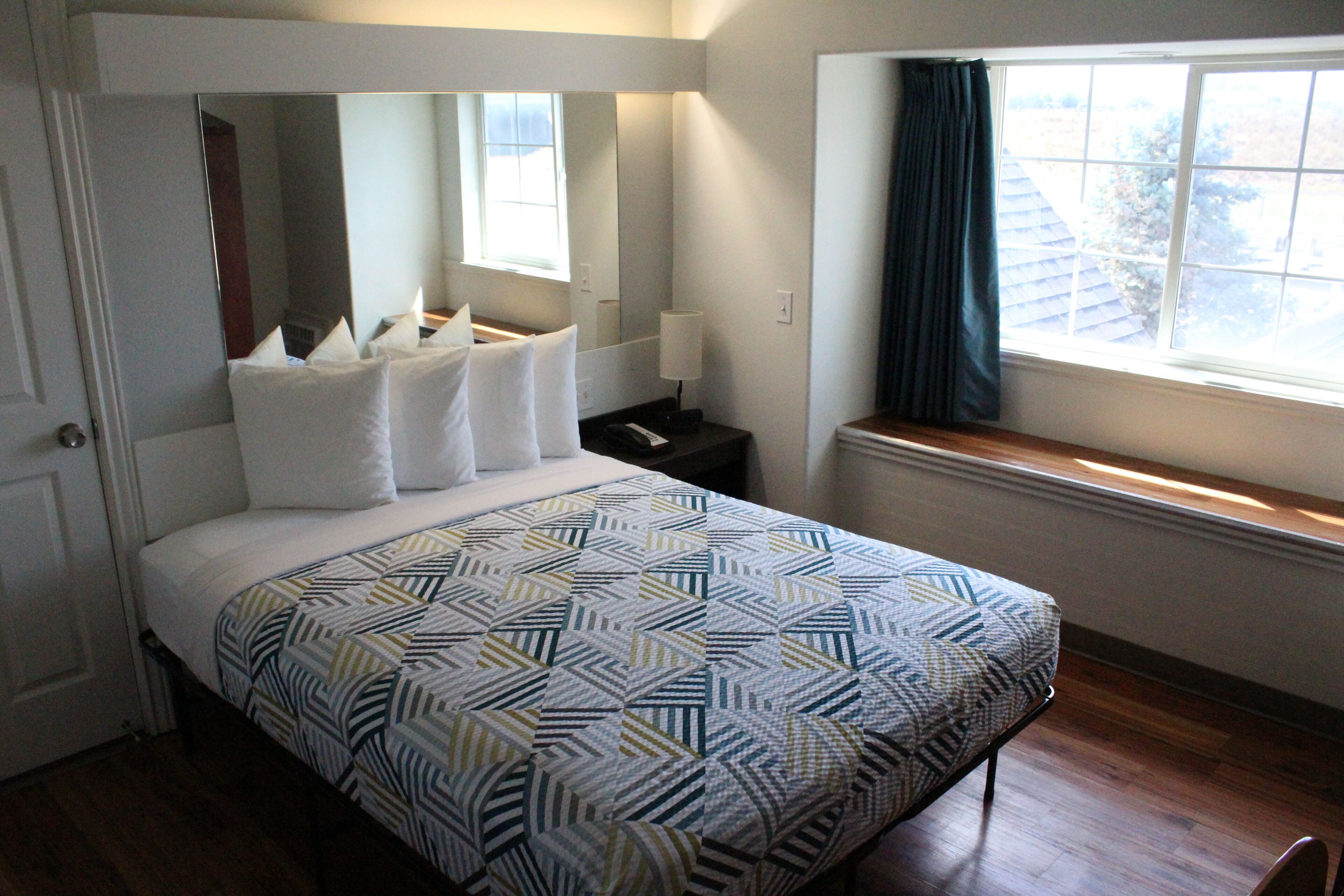Une chambre à coucher avec un lit, une table de chevet, un miroir, une fenêtre avec des rideaux et une vue sur l’extérieur.