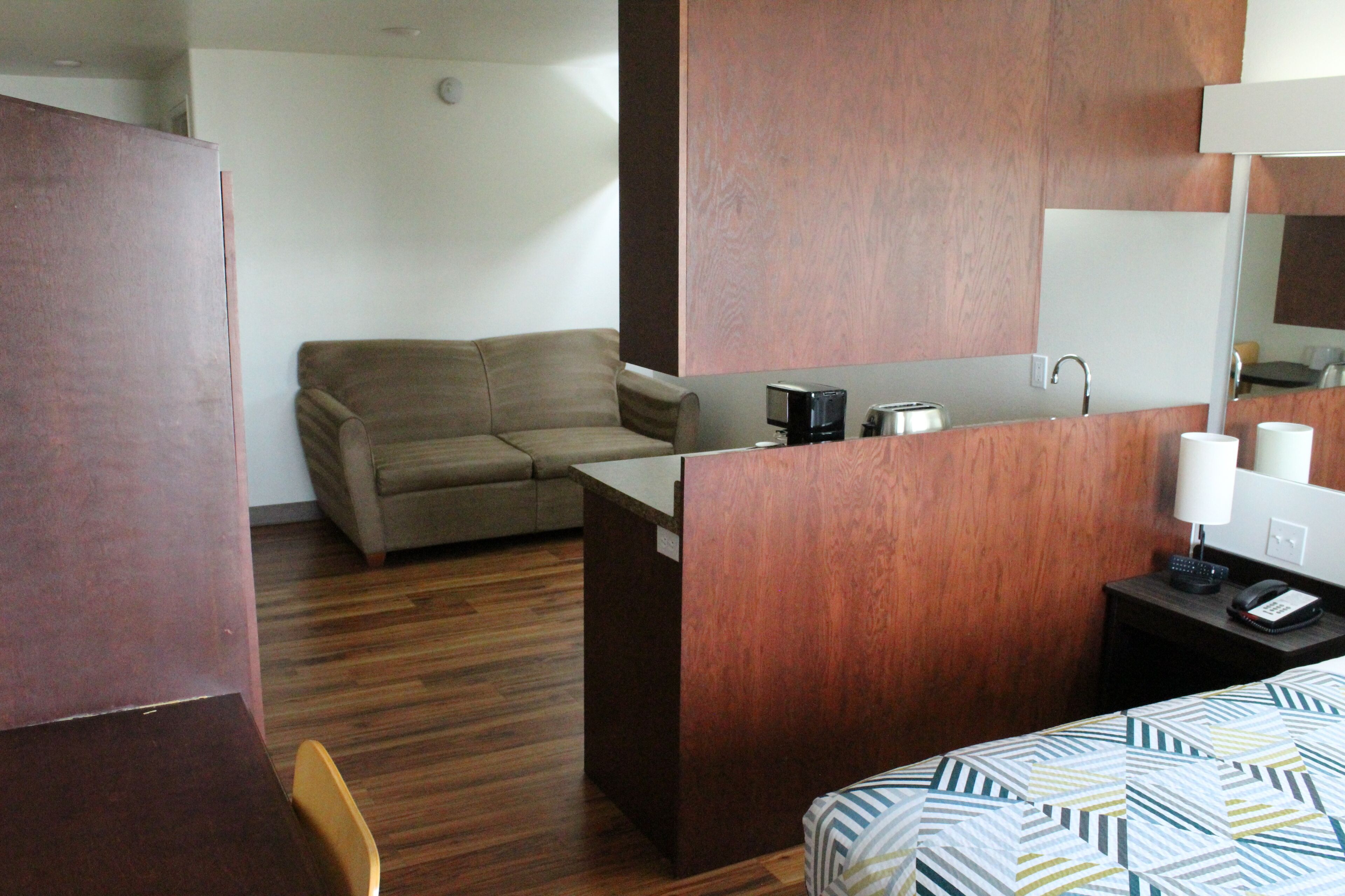 A hotel room with a wooden desk, a sofa, a bed with patterned bedding, a mirror, and a small kitchenette area.