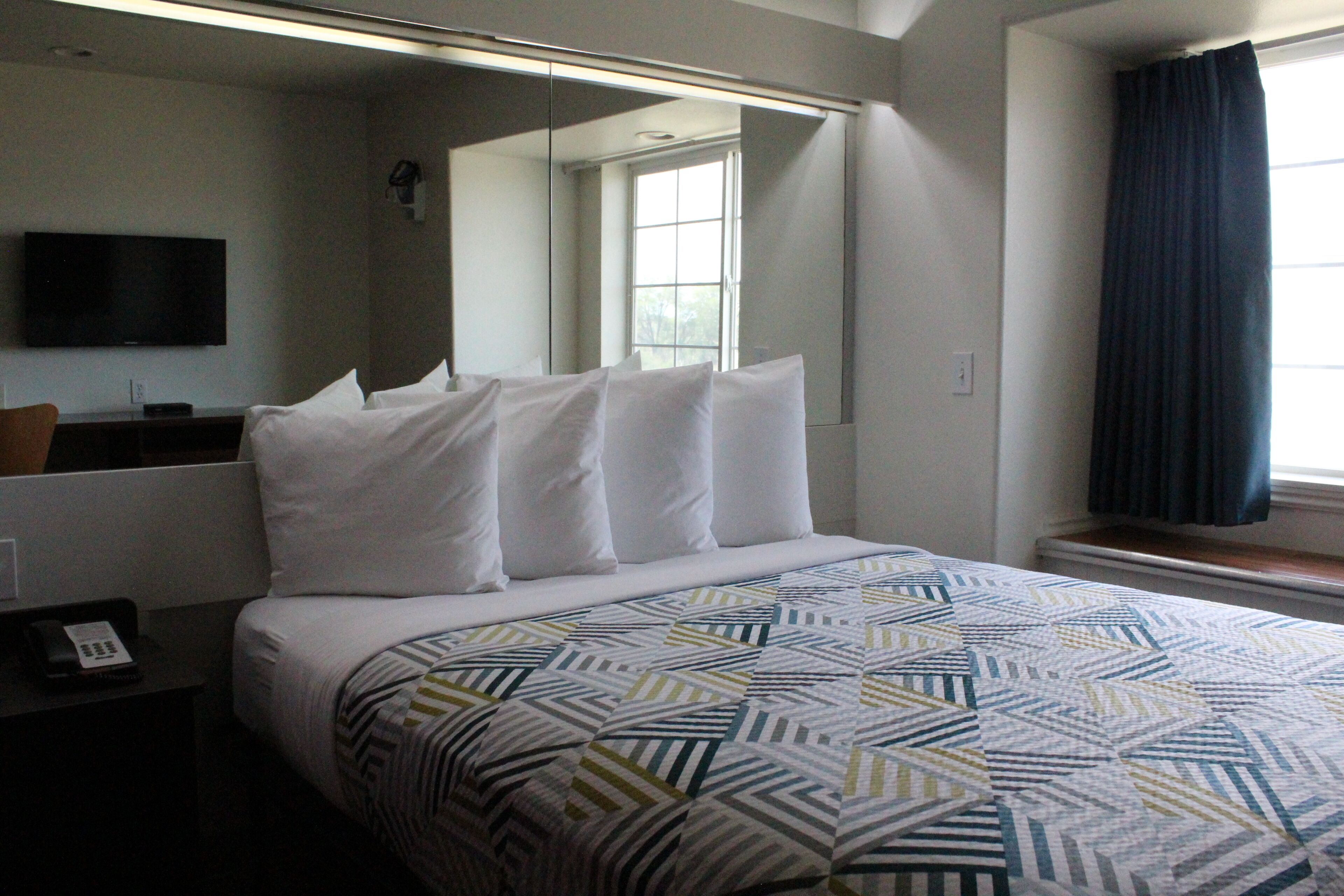 Una habitación de hotel con cama, televisor, ventana con cortinas y espejo.