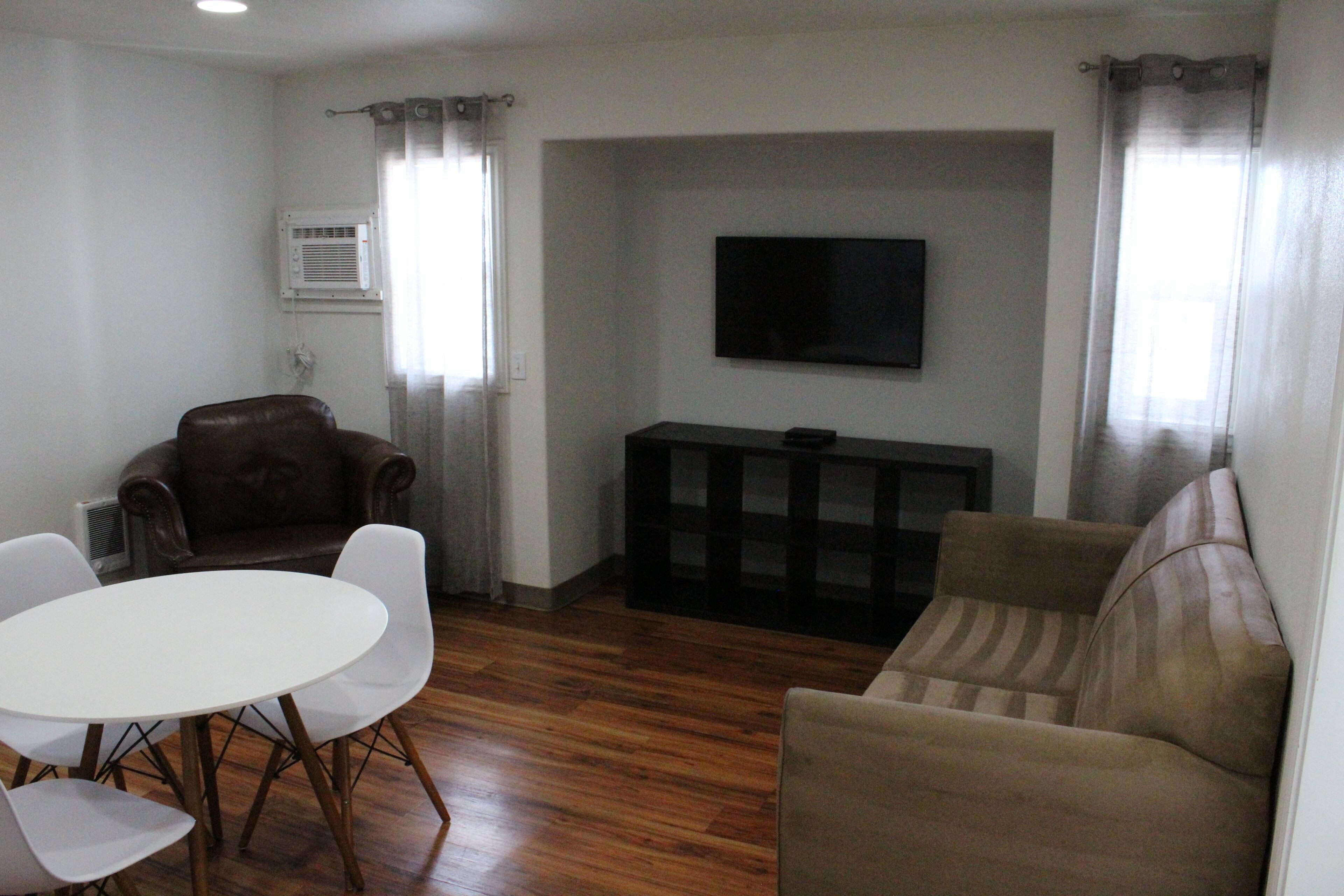 A living room with a white table, two chairs, a brown sofa, a TV mounted on the wall, and a window with curtains.