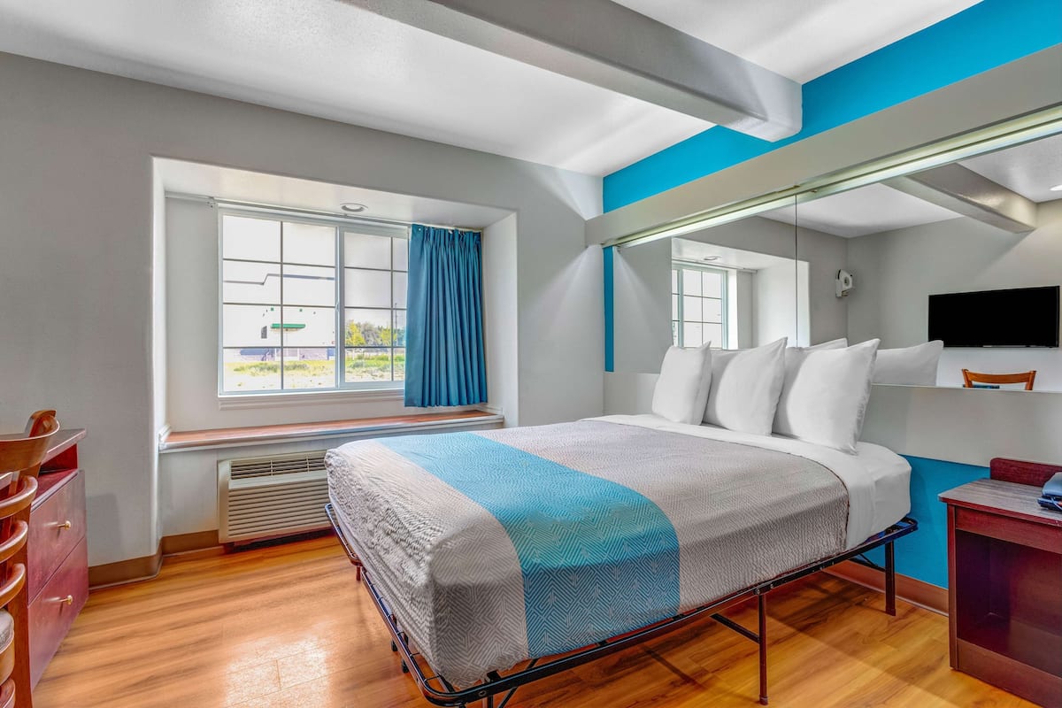 Ein Hotelzimmer mit Bett, Schreibtisch, Stuhl, einem Fenster mit Vorhängen und einem Fernseher an der Wand.