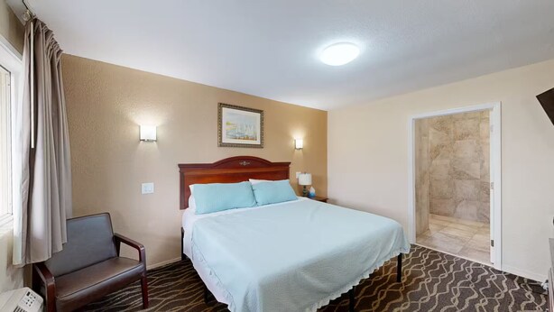 Ein Hotelzimmer mit Bett, Sessel, Schreibtisch, Lampe und einem Gemälde an der Wand.