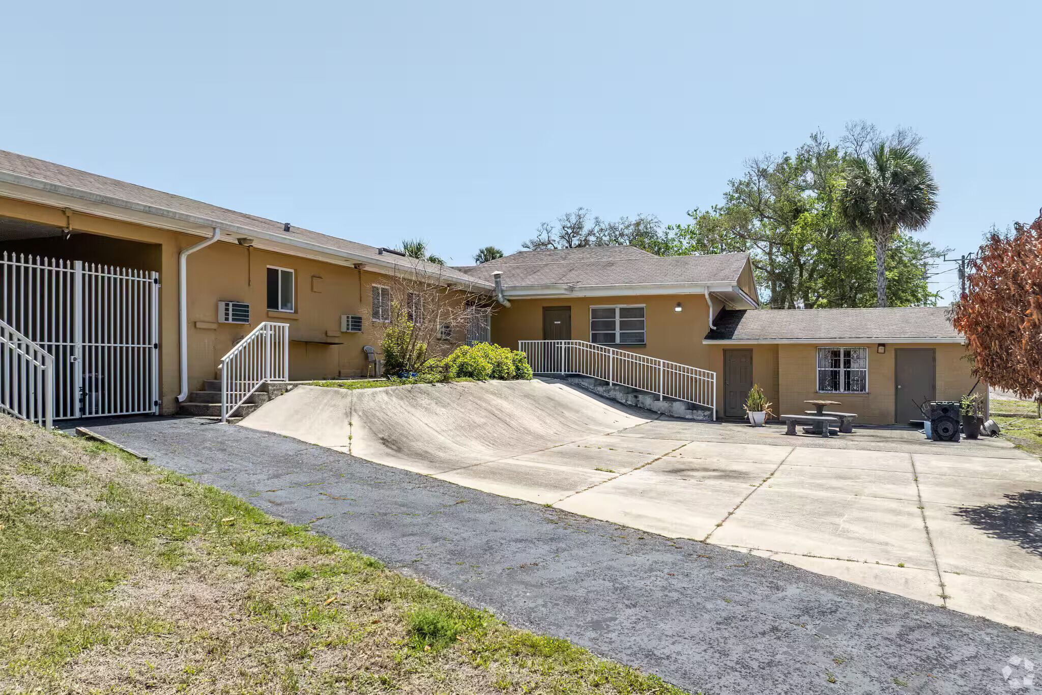 A building with a ramp leading to a covered area, a small patio with a table and chairs, and a fenced driveway.