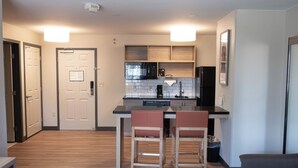 A modern kitchen with a black microwave, a coffee maker, and wooden cabinets. There are two bar stools and a trash can near the kitchen. - Candlewood Suites Sheridan by IHG (Sheridan)