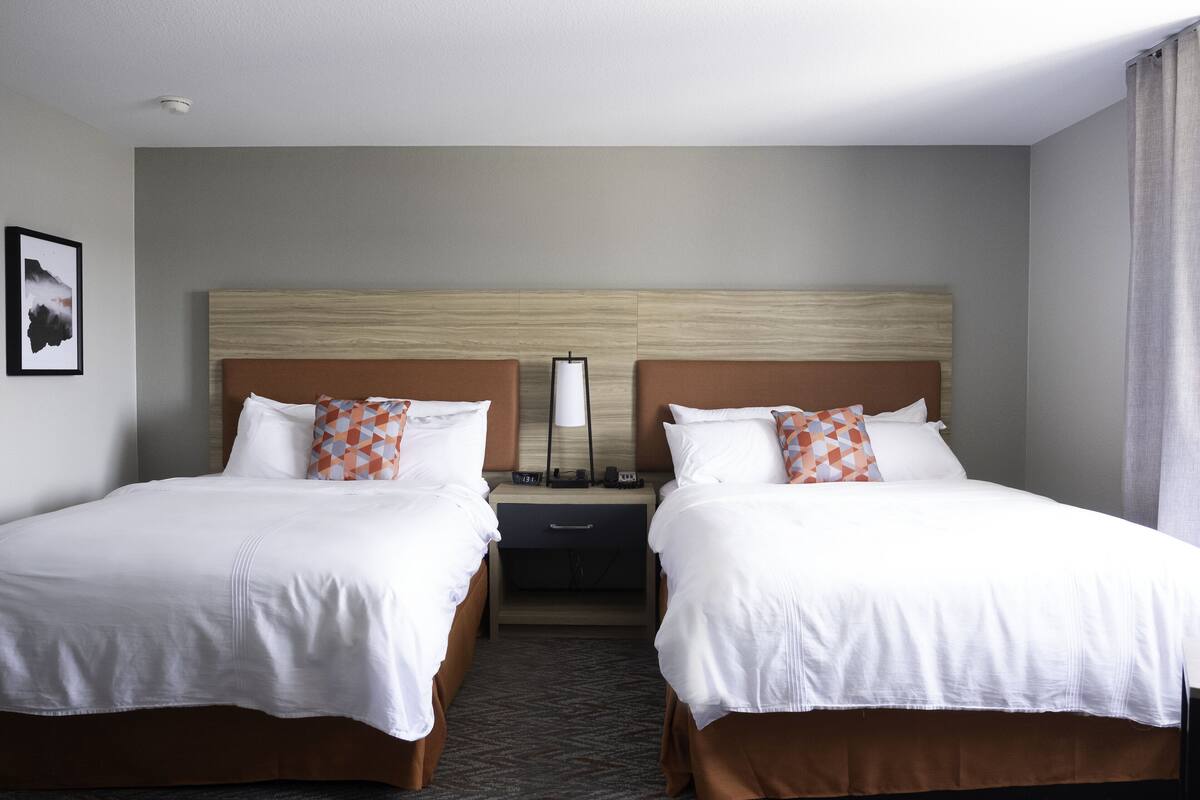 A hotel room with two beds, each with white bedding and brown headboards. There is a nightstand between the beds with a lamp and a clock. The room has a neutral color scheme with a gray wall and a framed picture on the left.