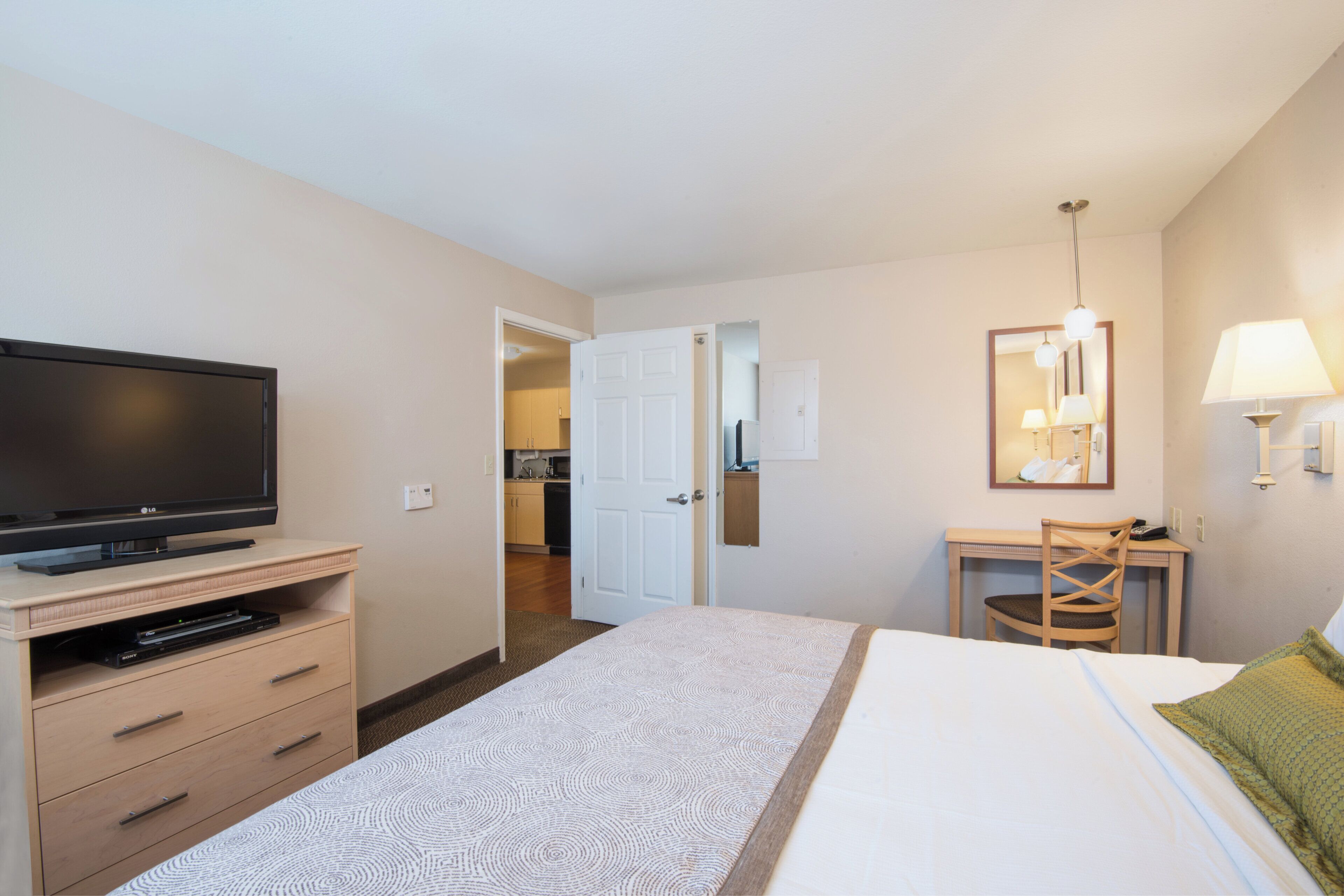 A hotel room with a bed, a desk with a chair, a television, and a kitchenette in the background.