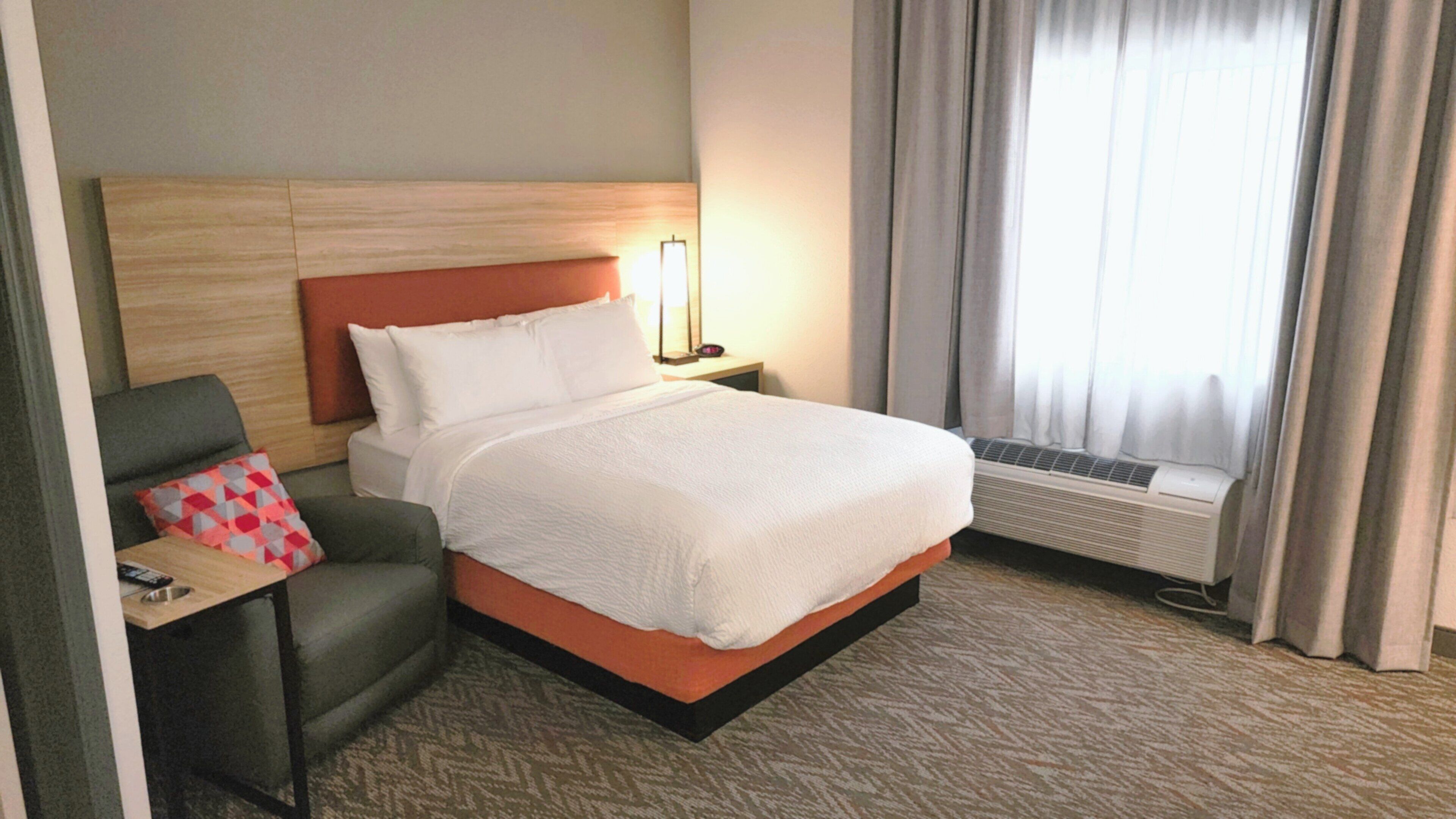 Une chambre d’hôtel comprenant un lit, une chaise, une table de chevet, une lampe, un appareil de climatisation et une fenêtre avec des rideaux.