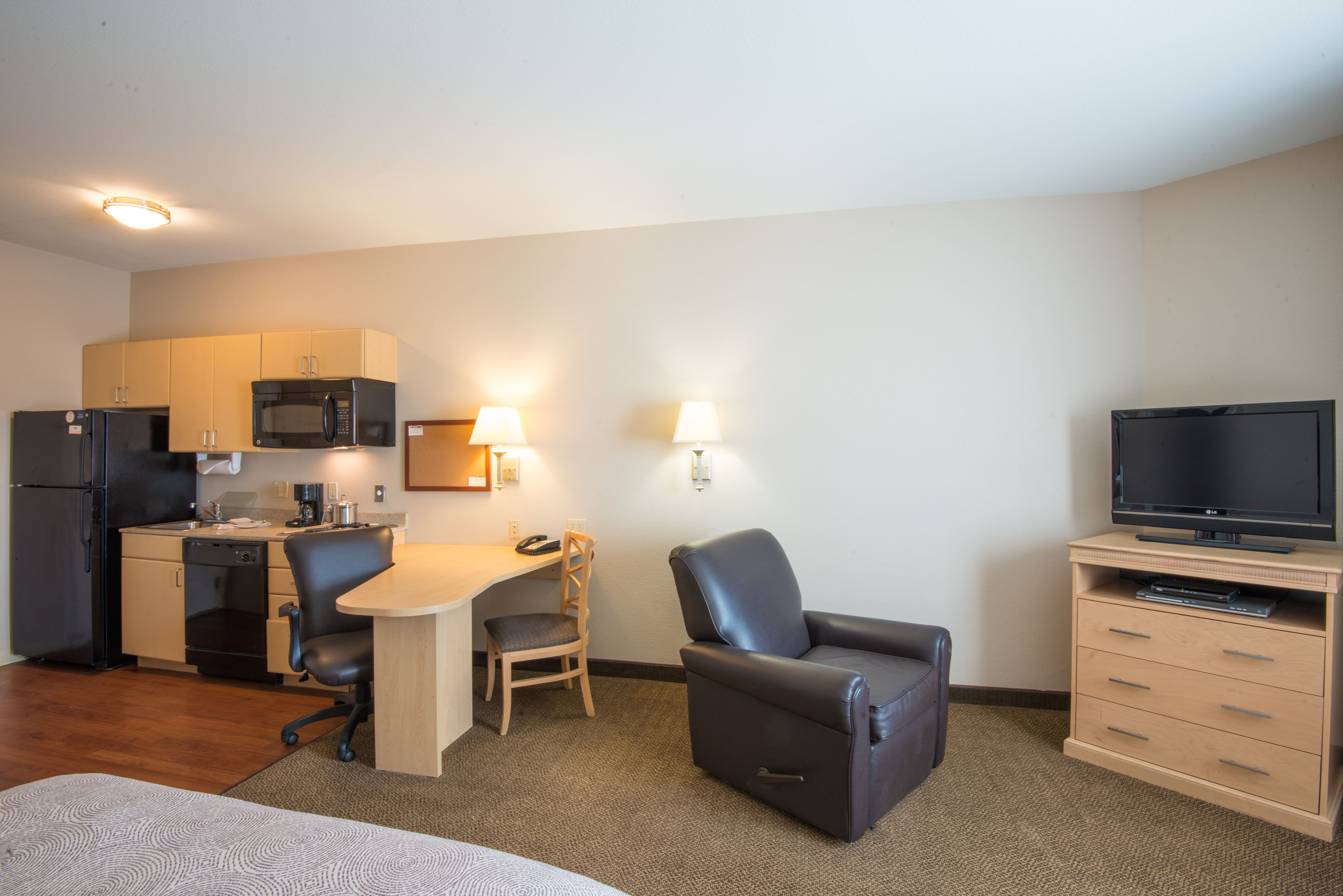 A hotel room with a kitchenette, a desk with a chair, a leather armchair, a TV, and a bed.
