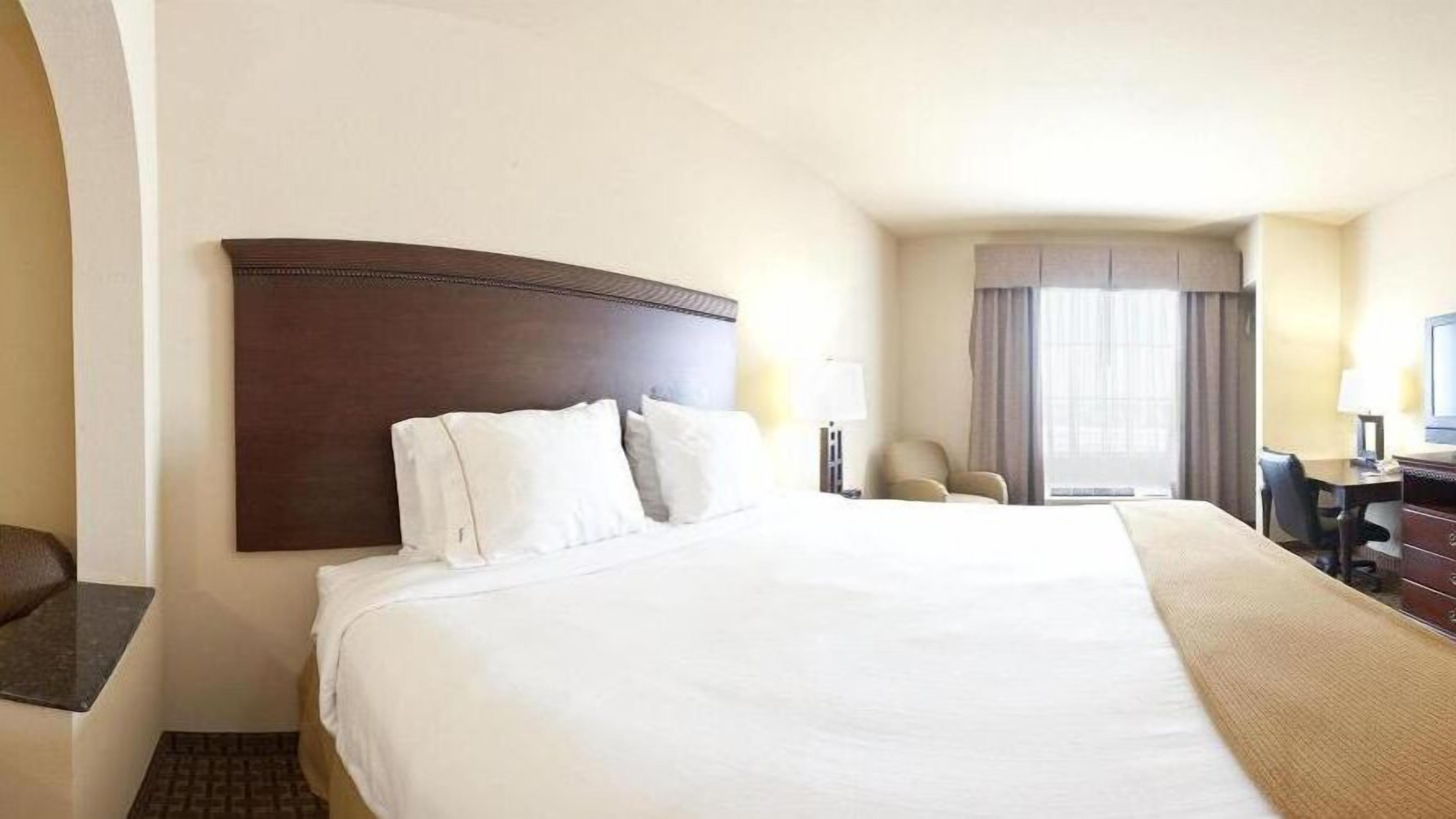 Quarto de hotel com uma cama grande, escrivaninha, cadeira e janela com cortinas.