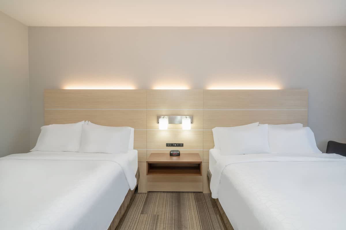 A hotel room with two beds, a wooden headboard, a nightstand with a clock, and a wall-mounted light fixture.