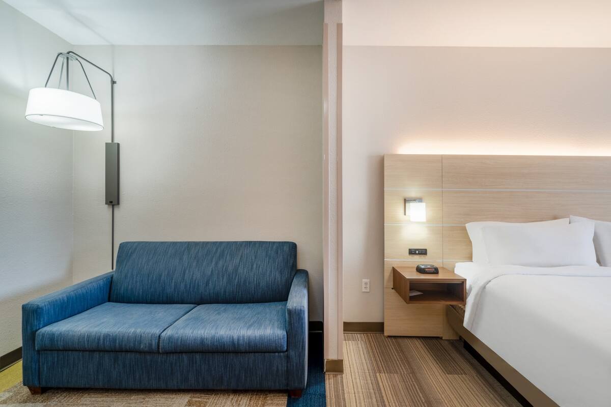 A hotel room with a blue sofa, a bed with white linens, a nightstand with a lamp, and a wooden headboard.