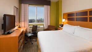 A hotel room with a bed, a desk with a television, a chair, a window with curtains, and a city view.